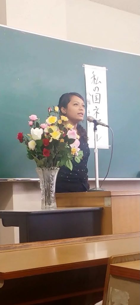黒板の前でマイクに向かい、「私の国 ネパール」と書かれた紙を背に発表するラジナさん