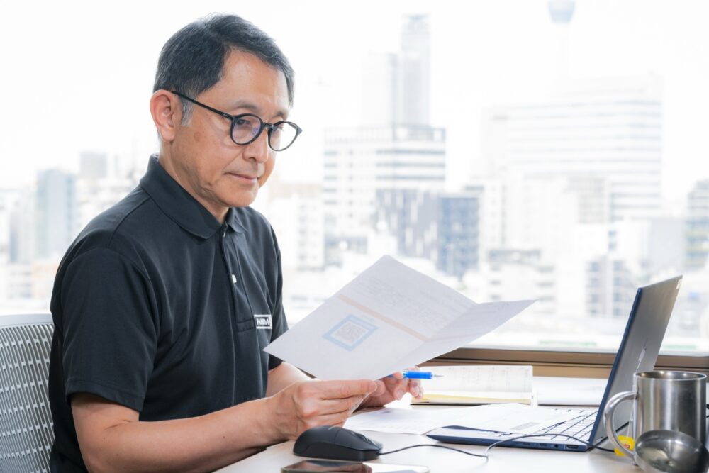 窓際のデスクで、書類を手に取って読む芥田さん