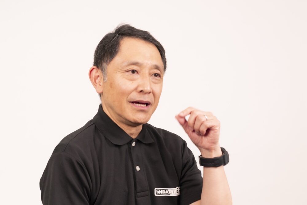 白い背景の前で、手を動かしながら話す芥田さん