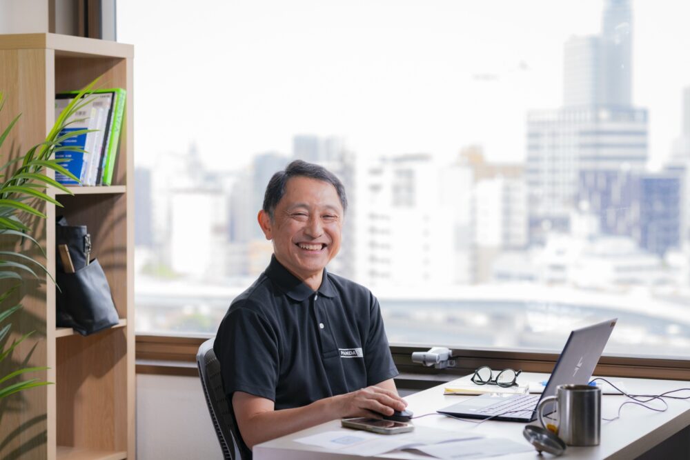 窓辺のデスクで、こちらを見て笑う芥田さん