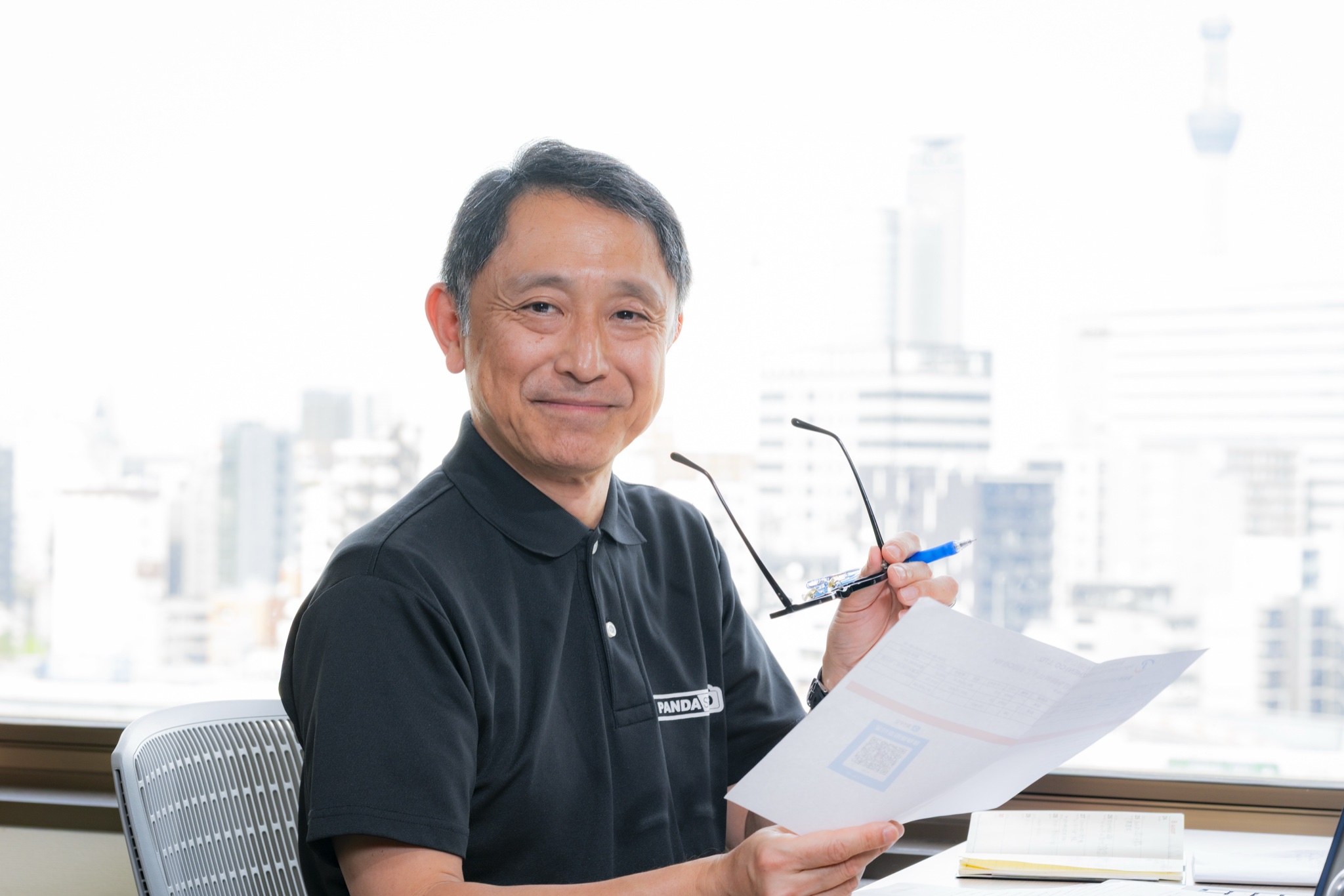 窓を背にしたデスクで、書類とメガネを手にこちらを見て笑う芥田雄輝さん